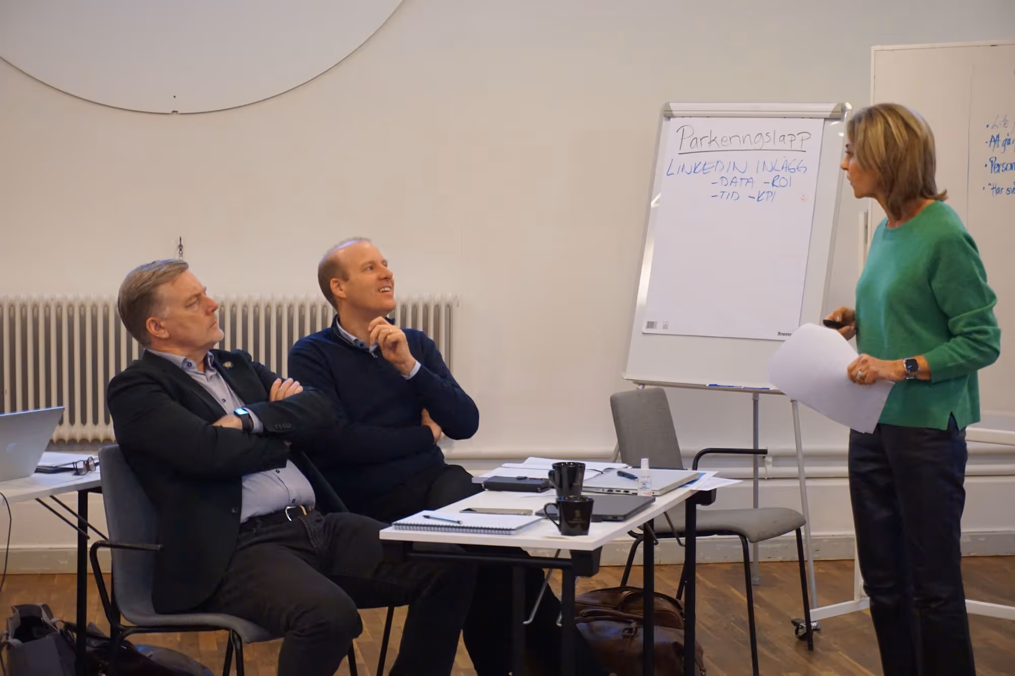 Två män sitter vid ett bord och lyssnar på en kvinna som presenterar med papper och whiteboard i ett mötesrum.