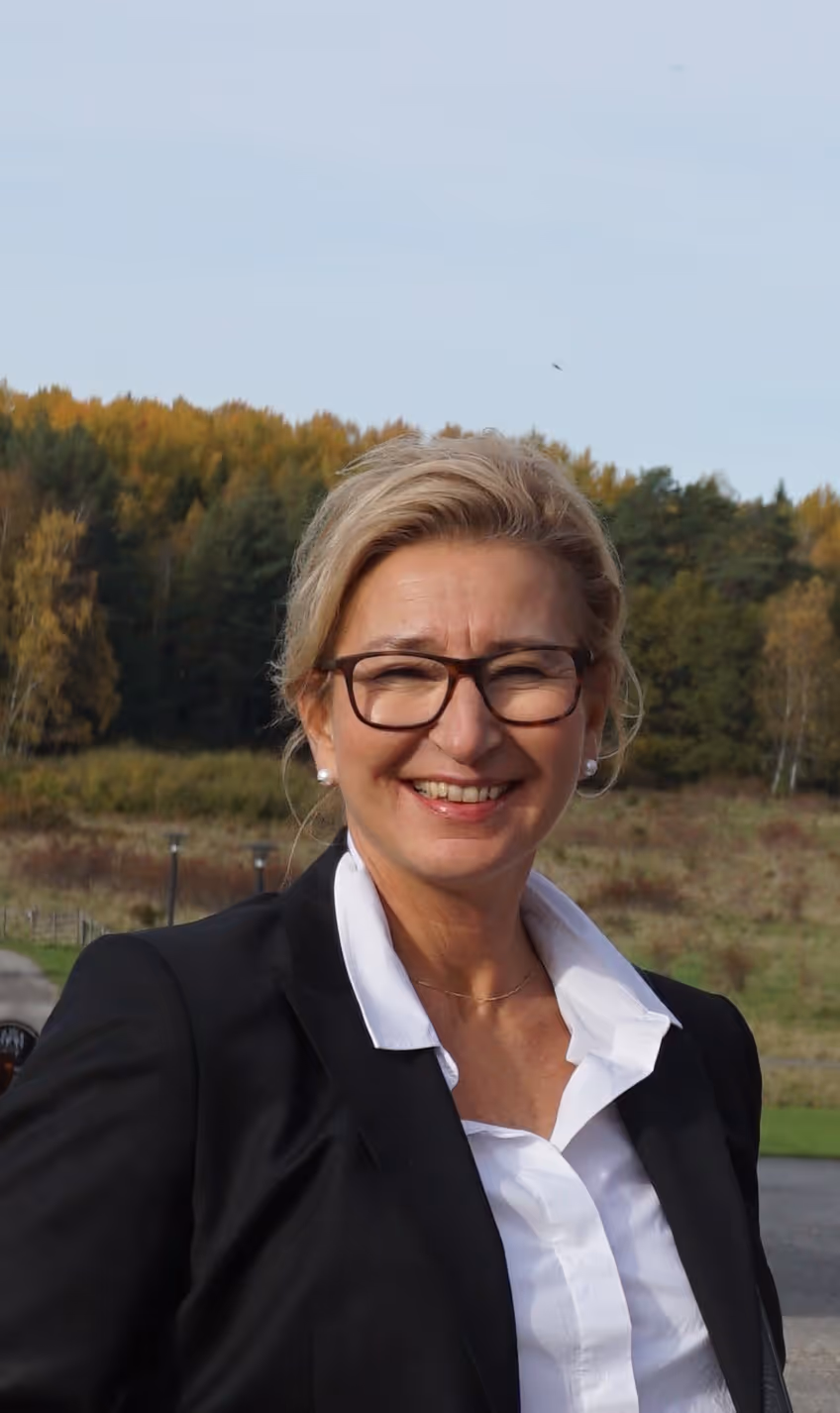 Kvinna med glasögon och svart kavaj ler utomhus med skog i bakgrunden.